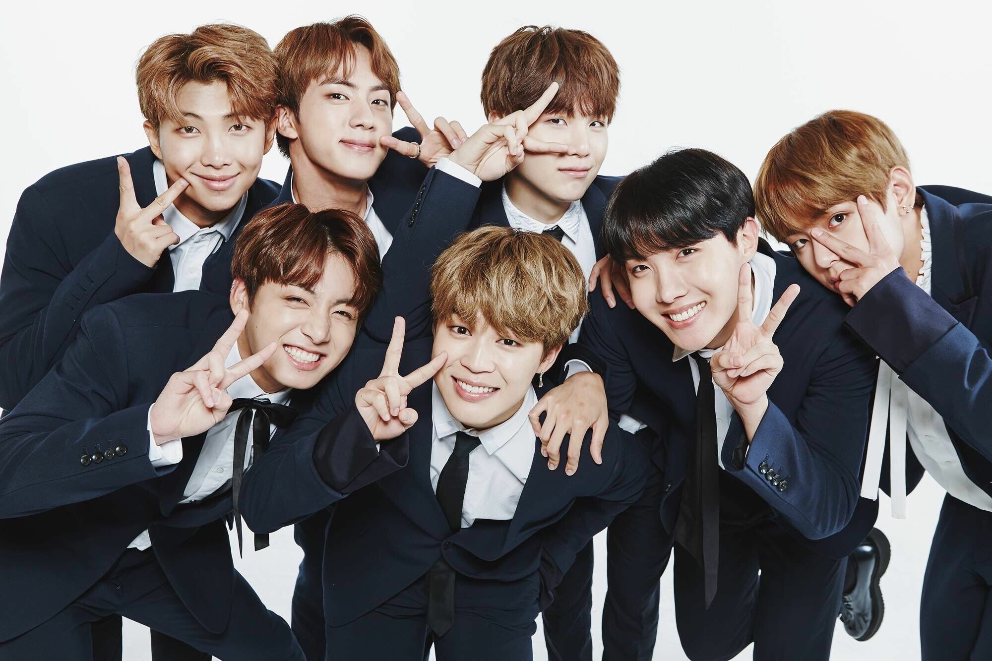 BTS members wearing matching navy blue suits, smiling and posing with peace signs.