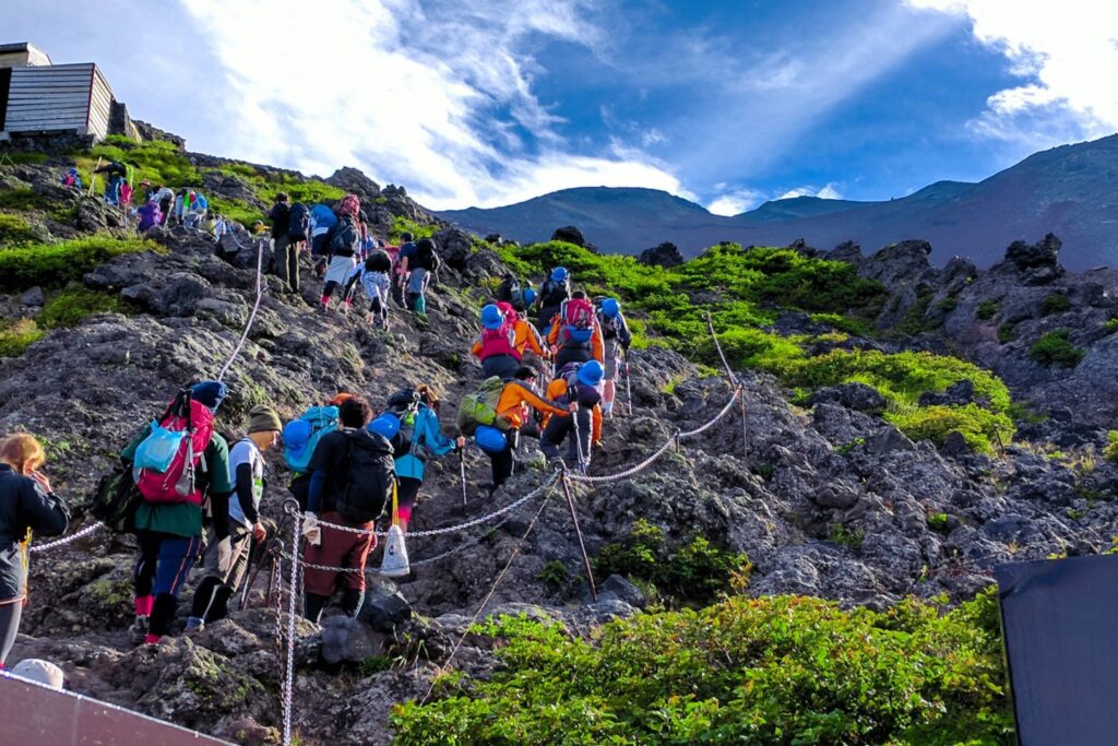 Hikers climbing the Yoshida Trail on Mount Fuji with volcanic rocks and shrines visible along the path.